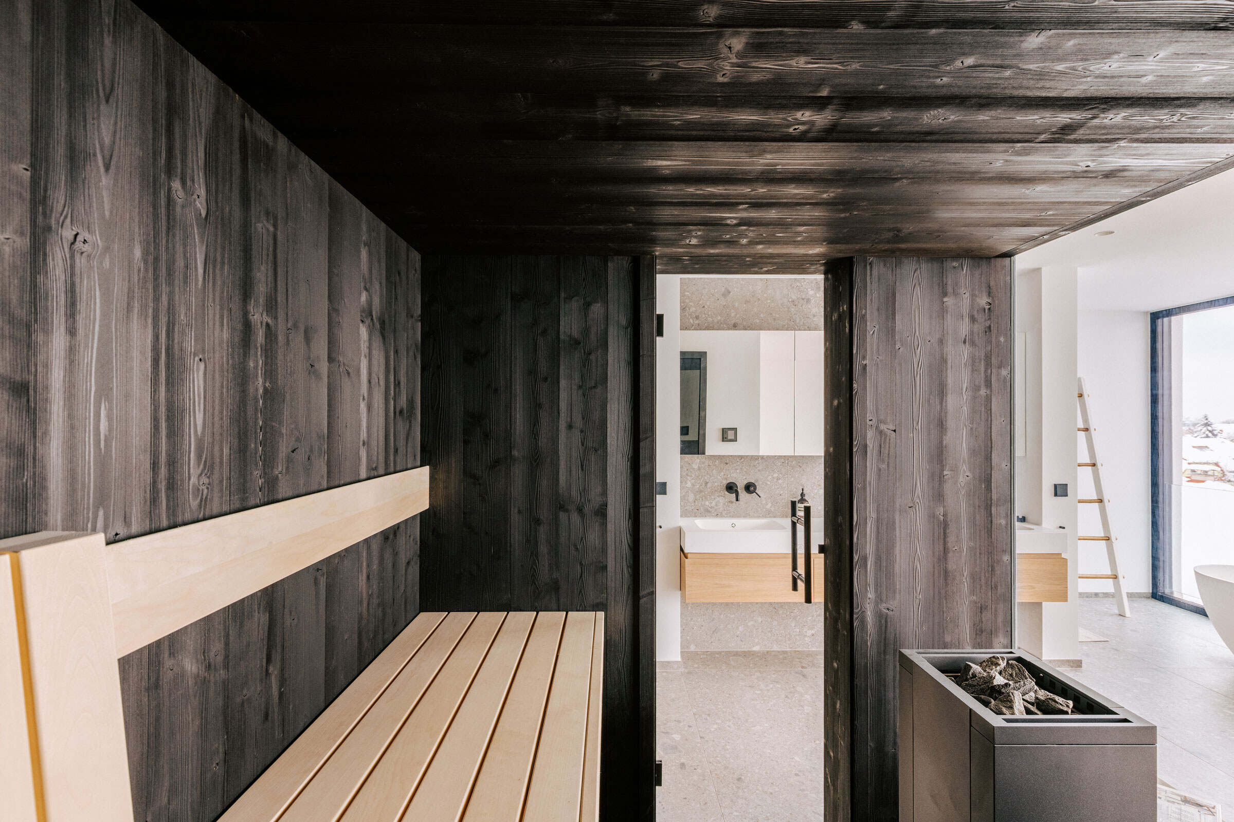 Luxus-Sauna nach Maß aus schwarzem Holz mit Verglasung und Blick in ein modernes Badezimmer mit Waschtisch