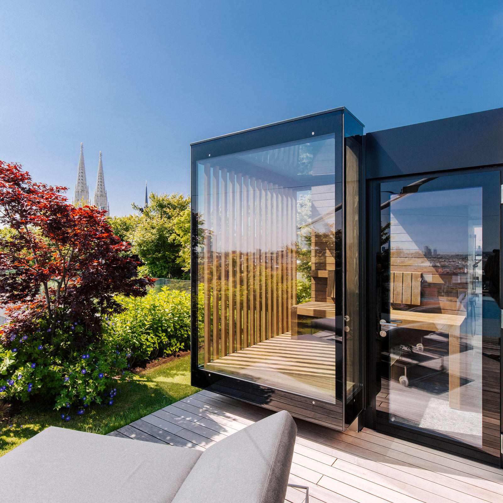 Voll verglaste Außensauna in modem Design mit seitlichem Sichtschutz auf einer begrünten Dachterrasse