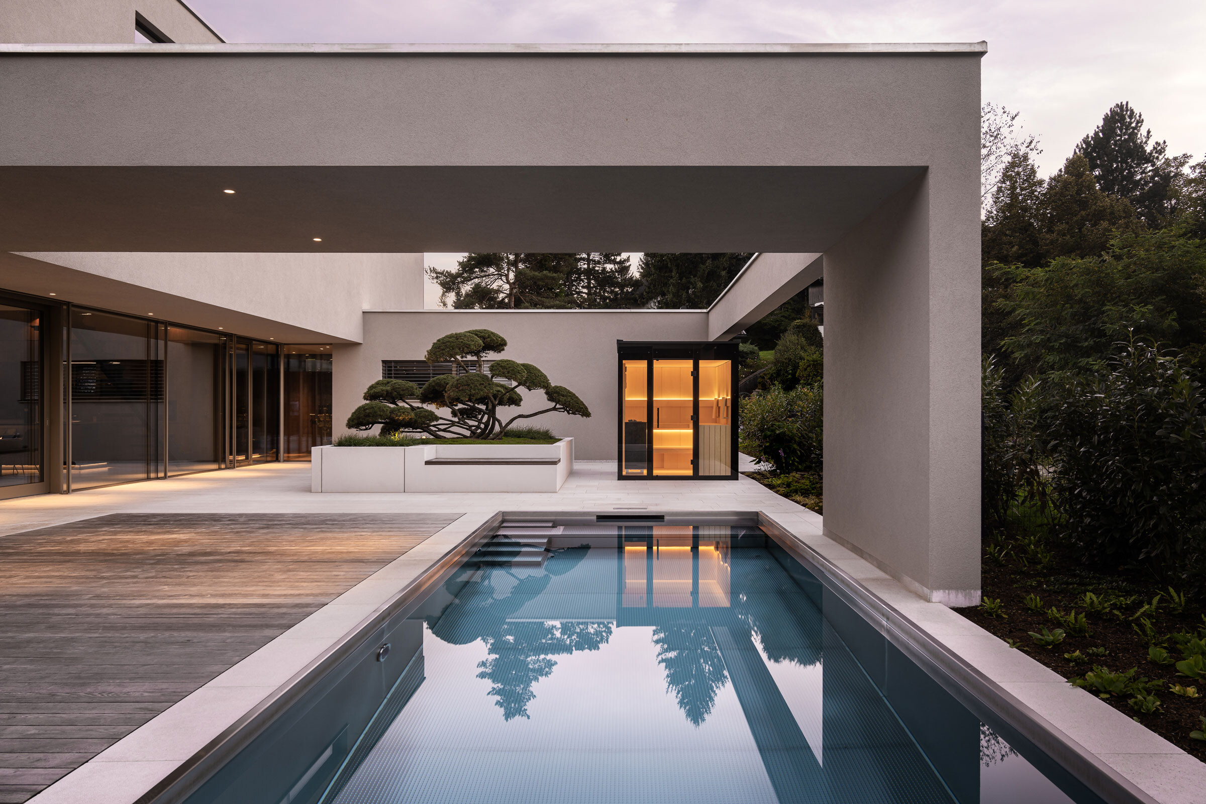 Modernes Architektenhaus mit beleuchteter Outdoor-Sauna auf einer Terrasse mit großem Pool in der Abenddämmerung