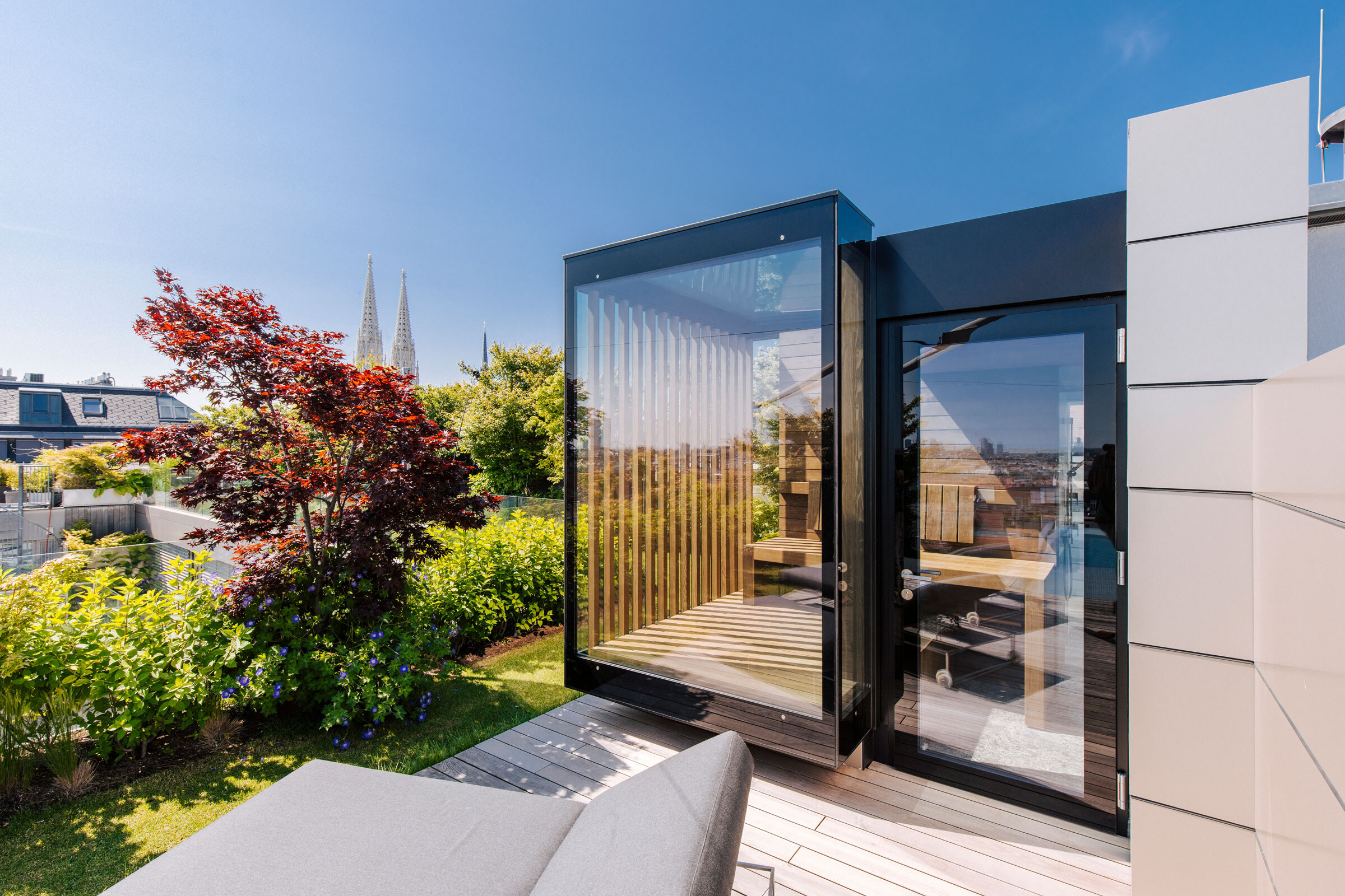 Verglaste Outdoor-Sauna von Deisl mit seitlichem Sichtschutz auf einer Dachterrasse mit Garten und schönem Ausblick